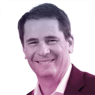 A man with short dark hair, wearing a suit jacket and collared shirt, smiling at the camera against a plain background.